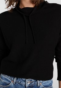 Svart huvtröja med hög krage, ribbad textur och dragsnören, i kombination med ljusblå denimjeans med knapplås.