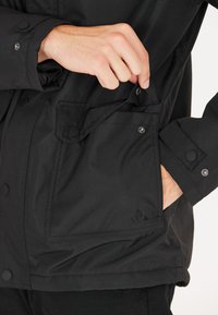 Zwarte jas van een gestructureerde stof, met drukknoopsluitingen en een zijzak. Subtiel logo op de rechter onderzijde.