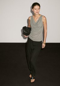 Gray draped sleeveless top, loose black wide-leg trousers, and black woven clutch. Model wearing flat black sandals, standing against a plain background.
