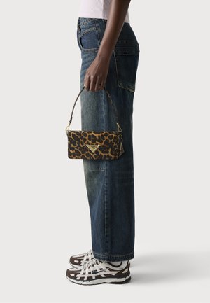 Sac à bandoulière en imprimé léopard en tissu, avec un logo triangulaire en or. Porté par une personne vêtue de jeans larges bleus et de baskets.