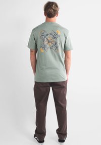 T-shirt vert menthe avec un design floral dans des teintes de gris, jaune et orange au dos, associé à un pantalon marron et des baskets noires.