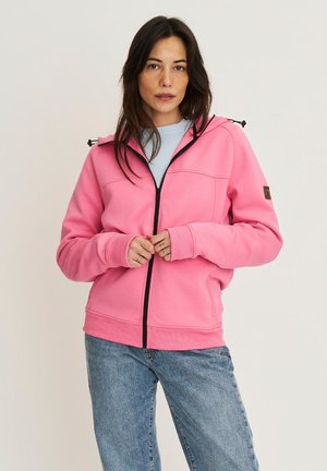 Vrouw met lang donker haar, gekleed in een roze hoodie met rits over een lichtblauw shirt en een blauwe spijkerbroek, staand tegen een effen achtergrond.