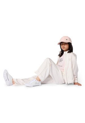 Veste blanche zippée portée sur un t-shirt rose clair, associée à un pantalon crème et des baskets blanches. Une casquette rose clair avec un logo complète le look.