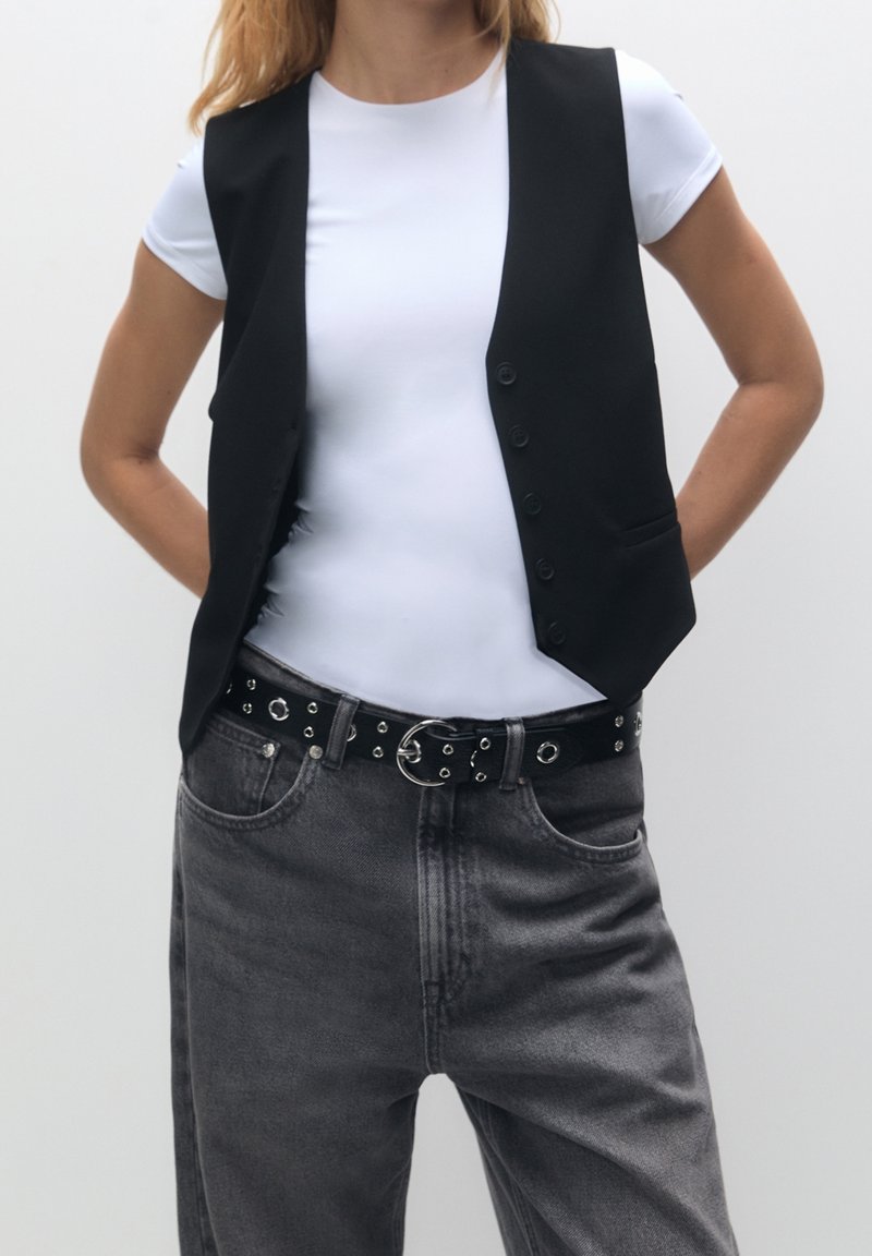 Personne portant un t-shirt blanc, un gilet noir boutonné, un jean gris avec une ceinture noire comportant des œillets métalliques et une boucle circulaire.