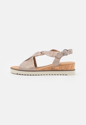 Tan kile-sandal med flettede remme, justerbar spænde, kork-mellemsål og hvid takkekantet gummisål på hvid baggrund.
