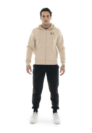Un hombre luce una sudadera con capucha beige con cremallera, pantalones deportivos negros y zapatillas blancas sobre un fondo blanco liso.