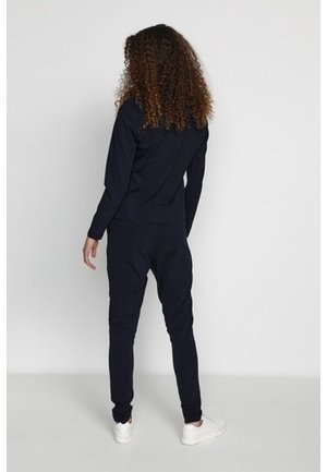 Personne aux cheveux bouclés vue de dos, portant un survêtement noir et des baskets blanches sur un fond clair uni.