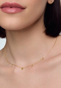 Zbliżenie na szyję modelki, eksponujące delikatny złoty naszyjnik z małymi zwisającymi kółkami, uzupełniający jej gładką skórę i subtelny makijaż.
