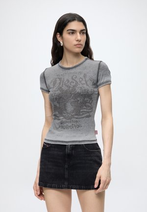 Donna con capelli lunghi e scuri che indossa una maglietta grigia sbiadita con grafica e una minigonna in denim nera, in piedi davanti a uno sfondo chiaro.