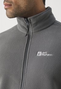 Szara polarowa kurtka z wysokim kołnierzem, zamkiem błyskawicznym z przodu oraz logo Jack Wolfskin haftowanym na białym tle na klatce piersiowej. Miękka tekstura i casualowy design.