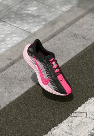 Schwarzer und pinker Nike ZoomX Laufschuh auf Asphalt in der Nähe eines Bordsteins mit taktilem Belag und einer weiß gestrichenen Linie.