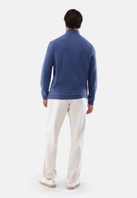 Blauer Strickpullover mit hohem Kragen, strukturiertem Stoff und gerippten Bündchen, kombiniert mit weißen Hosen und hellen, lässigen Schuhen.