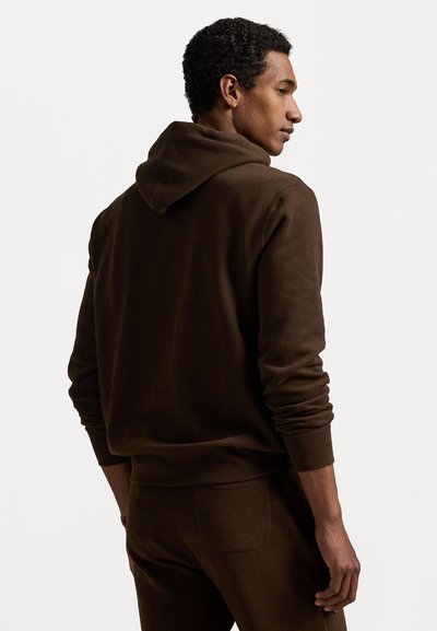 Rjava kapuca s kapuco, izdelana iz mehkega blaga, z ribastimi manšetami in pasom. Gladka tekstura, sproščen kroj in podrobno šivanje.