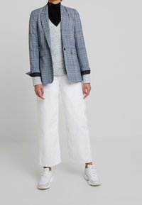 Blauer kariertes Sakko mit schwarzen Akzenten, grauer Pullover darunter, weite weiße Hose und graue Turnschuhe. Strukturierte Stoffe und maßgeschneiderte Passform.