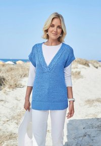 Lichtblauwe gebreide tuniek met een diepe v-hals en geribbelde zoom, gedragen over een wit longsleeve shirt. Ontspannen strandomgeving op de achtergrond.