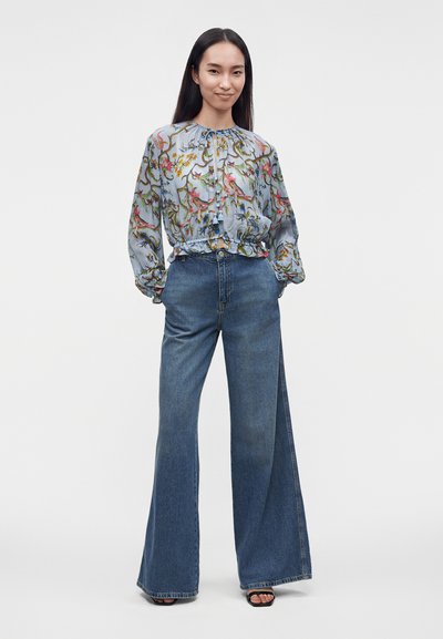 Femme debout, portant un chemisier bleu clair à fleurs à manches longues et un jean bleu taille haute à jambes larges, mains dans les poches, fond neutre.