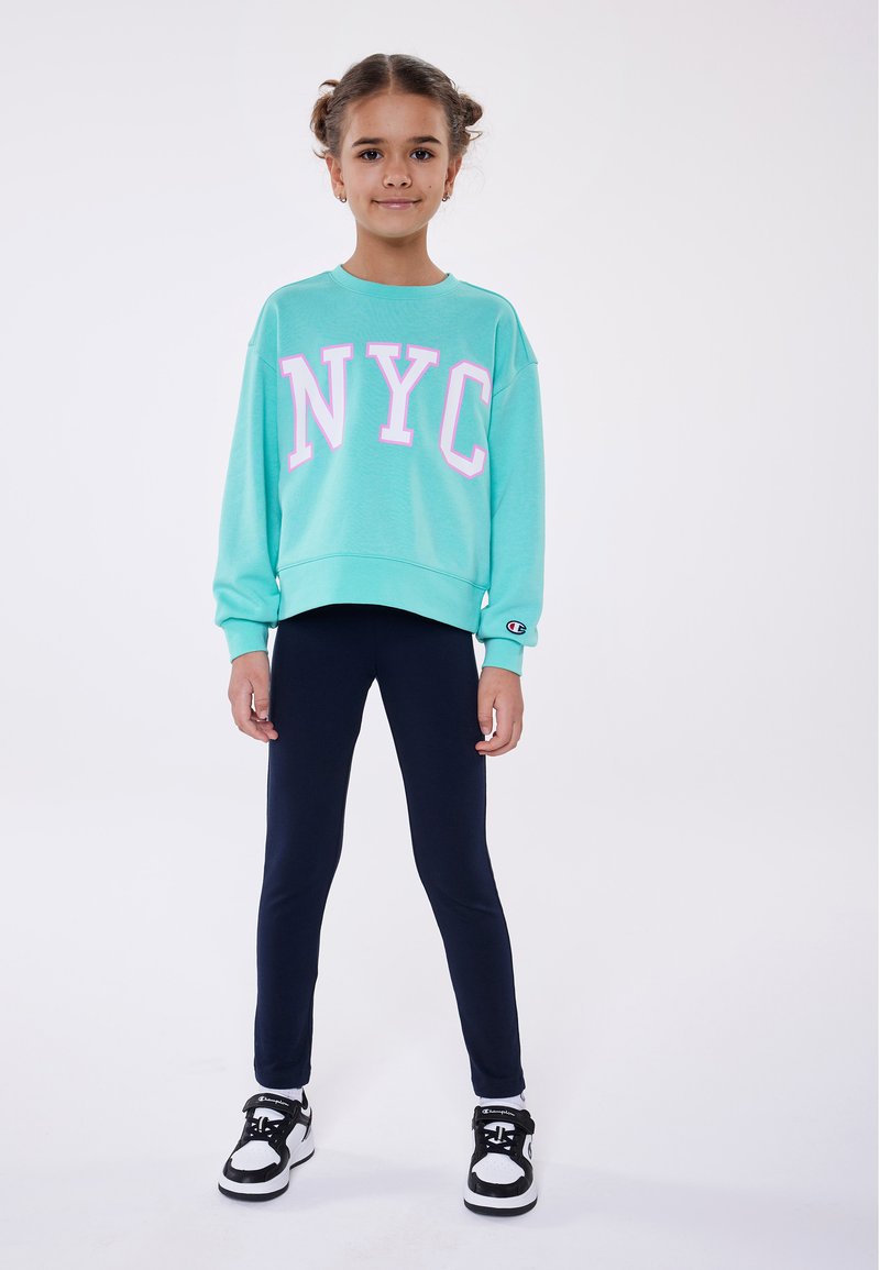 Felpa verde menta con "NYC" in lettere rosa evidenti, abbinata a leggings blu navy e scarpe da ginnastica bianco e nero. Leggera, dal taglio rilassato.