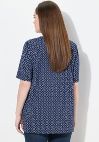 Námorně modrý top s krátkými rukávy, zdobený geometrickým světle modrým vzorem, kulatým výstřihem a volným střihem. Hladká textura s delším zadním dílem.
