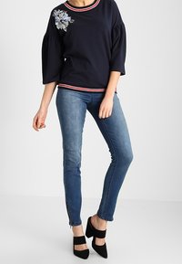 Maglione navy con maniche a sbuffo, scollo a righe rosse e bianche, ricamo floreale sulla spalla sinistra, abbinato a jeans blu slim e tacchi neri.