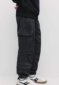 Pantaloni cargo negri cu multiple buzunare, manșete elastice și o croială lejeră, asortați cu adidași albi cu accente negre și bej.