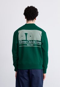 Sudadera de algodón verde con un diseño gráfico blanco que presenta líneas curvas y el texto 'Ellesse Club di Corsa' en la parte trasera. Textura suave.
