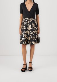Svart omslagsklänning med korta ärmar, prydd med ett mönster av gyllene bladblommor på kjolen, i kombination med svarta remsandaler med plattformsklack.