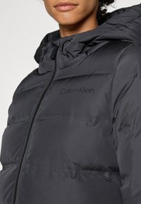 Čierna puffrová bunda so zipsom vpredu, nastaviteľnou kapucňou a logom Calvin Klein. Má hladkú, matnú textúru a prešitie.