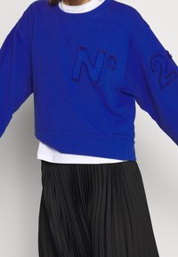 Persona con un sudadera azul con el bordado negro "N°2" en el frente y la manga, superpuesta a una camisa blanca, acompañada de una falda negra plisada.