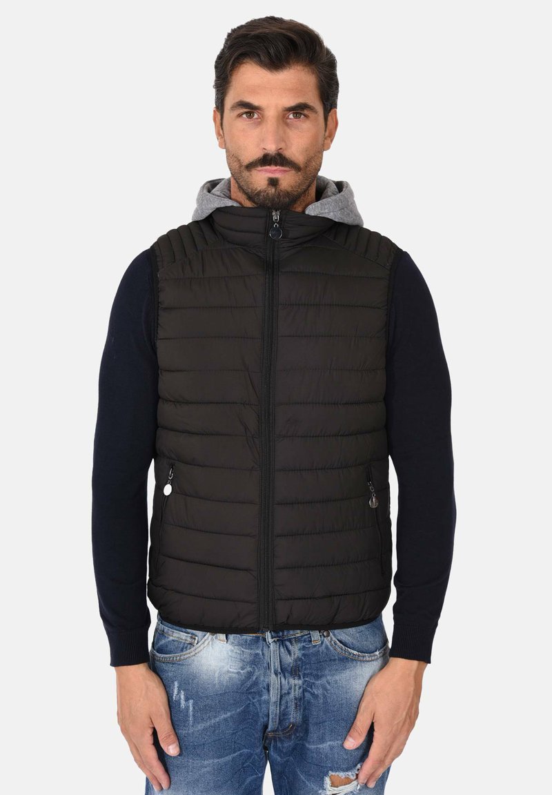 Gilet imbottito nero con cuciture orizzontali, chiusura con zip e cappuccio grigio. Indossato sopra una maglietta a maniche lunghe blu navy, abbinato a jeans blu chiaro sbiaditi.