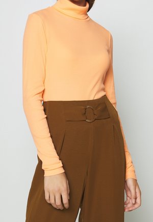 Personne portant un col roulé pêche à manches longues rentrée dans un pantalon marron taille haute avec une large ceinture et une boucle circulaire.