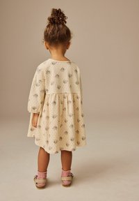 Kleid mit floralen Mustern in crèmefarbe, mit Puffärmeln und einem gerafften Rock. Das Kind trägt rosa strukturierte Socken und rosa Sandalen mit einer Schnalle.