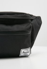 Sac banane en tissu noir avec poche zippée à l’avant et étiquette blanche rectangulaire de la marque Herschel en bas à droite.