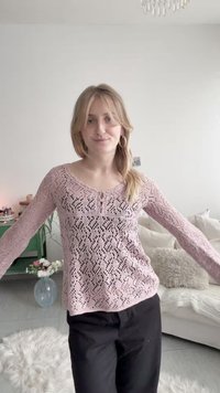 Νεαρή γυναίκα που φορεί ένα ανοιχτό ροζ πλεκτό μακρυμάνικο τοπ και μαύρο παντελόνι, στέκεται σε ένα φωτεινό σαλόνι με λευκό καναπέ και λουλούδια.