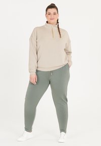 Beige zip-up sweatshirt med långa ärmar, parat med ljusgröna joggers. Båda plagg är tillverkade av mjukt tyg. Vita sneakers fulländar outfiten.