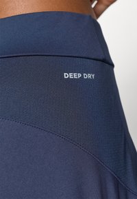 Közelkép sötétkék sportleggingsről, textúrált anyagpanellel és a deréknál elhelyezett "DEEP DRY" logóval.