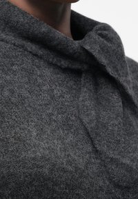Vêtement en laine texturé gris foncé avec un col montant et un détail de lien en tissu autour du cou, porté par une personne.