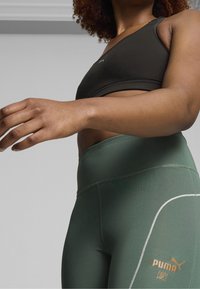 Mustat urheiluliivit, joissa on tyylikäs muotoilu, yhdistettynä korkeavyötäröisiin tummanvihreisiin leggings-housuihin, joissa on valkoinen yksityiskohta ja logo.