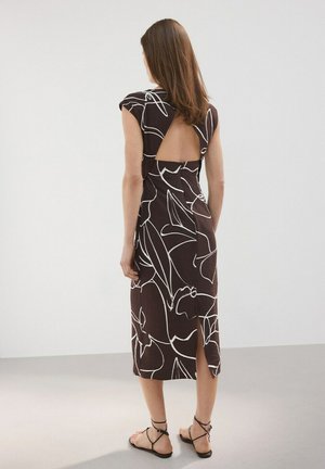 Kobieta w brązowej sukience do kolan z białymi abstrakcyjnymi kwiatowymi liniami i trójkątnym, odkrytym tyłem, stojąca na jasnoszarej podłodze.