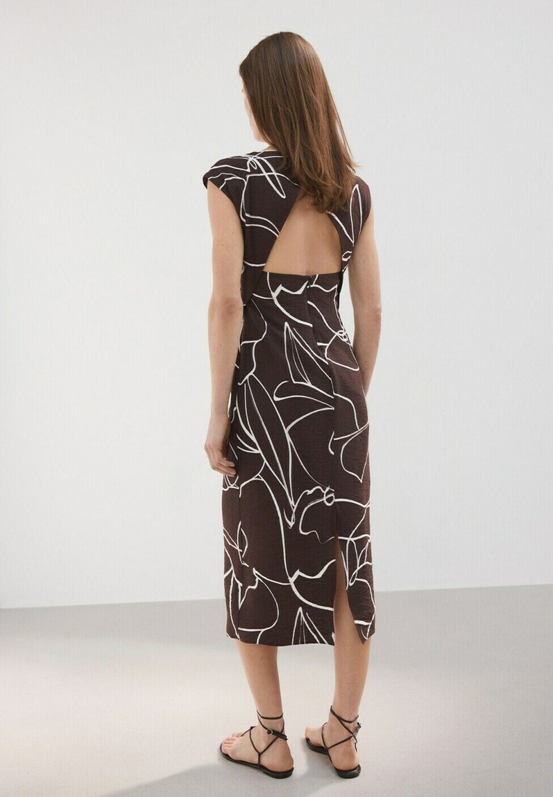 Femme portant une robe marron longueur genou avec des lignes florales abstraites blanches et un dos ouvert en forme de triangle, debout sur un sol gris clair.