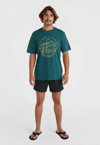 Aquafarvet bomulds t-shirt med rund halsudskæring og korte ærmer, prydet med en gul grafik, der viser bølger og sol. Båret med sorte shorts og flip-flops.