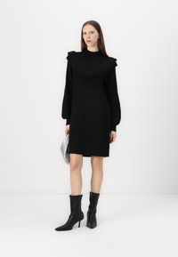 Juoda megzta suknelė su V formos apykakle, ilgomis rankovėmis ir raštuota tekstūra. Dereita su juodais smailėjančiais auliniais batais ir sidabrine rankine.