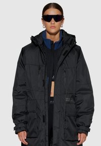 Czarna kurtka puchowa z kapturem, zapięciem na zamek i wieloma kieszeniami. Posiada niebieskie akcenty i teksturowane wykończenie. Nosi się ją na czarną bluzkę.