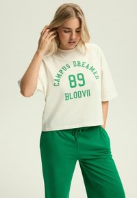 Kremowa, krótkotali t-shirt z zielonym nadrukiem "CAMPUS DREAMER 89 BLOOVII". W połączeniu ze zielonymi spodniami dresowymi z regulowanym paskiem. Gładki materiał, luźny krój.
