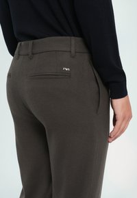 Tamsiai žali kelnės iš tekstūruoto audinio, turinčios galinius kišenes ir mažą metalinę logotipą dešinėje galinėje kišenėje.