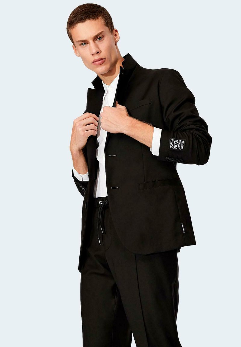 Blazer negro con un diseño estructurado, acentos plateados y detalles de puño abotonado. Usado sobre una camisa blanca, con un cuello alto.