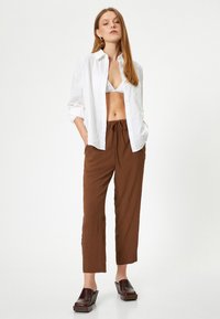 Witte linnen blouse met knopen, bruine textuur broek en bruine blokhak schoenen; de outfit heeft een ontspannen pasvorm en een overwegend neutraal kleurenpalet.