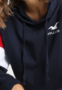Mörkblå hoodie med vit text "HOLLISTER" och en logotyp. Har röda och vita randiga detaljer på ärmarna och snoddarna. Mjukt tyg.