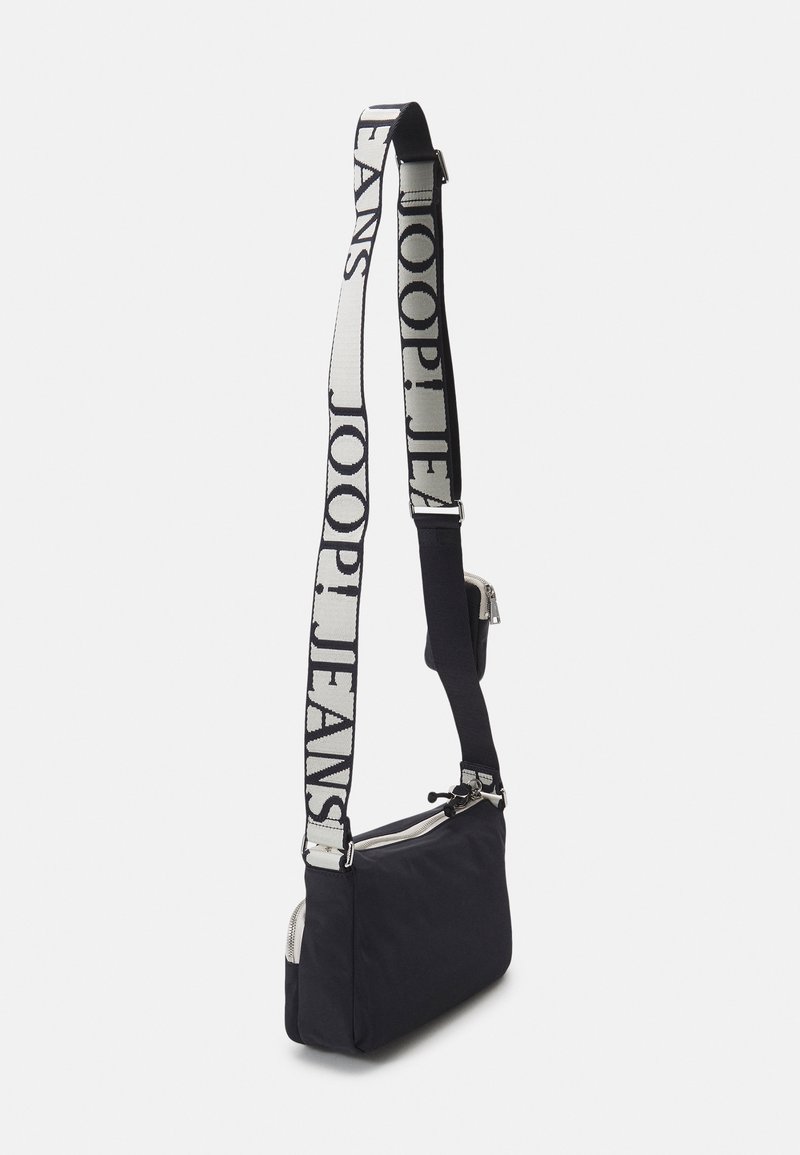 Schwarze Nylontasche zum Crossbody-Tragen mit Reißverschluss, ausgestattet mit einem breiten Trageriemen, auf dem "JOOP!JEANS" in fetten weißen Buchstaben gedruckt ist. Glatte Textur.