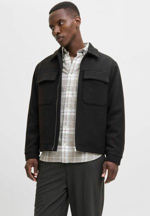 Homme portant une veste noire zippée par-dessus une chemise à carreaux beige et un pantalon foncé, debout sur un fond clair uni, regardant vers la gauche.