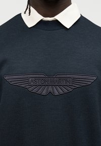 Σκούρα μπλε φούτερ με λευκό γιακά και ανάγλυφο λογότυπο Aston Martin σε γκρι στο στήθος. Μαλακή, λεία υφή.